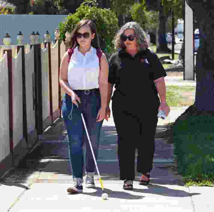 A young adult walking down the street using their cane while a Guide Dog staff member walks beside them.