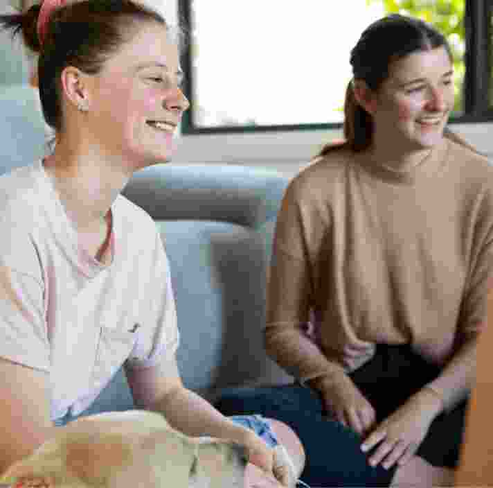 Three young adults sitting inside with a yellow labrador dog next to one of them. They are all looking at each other smiling.