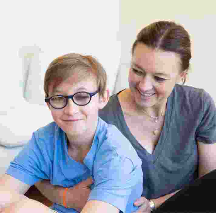 A young child with their mother seated on the ground. The mother is holding the child from behind and looking at her child. The child is looking at the camera smiling.